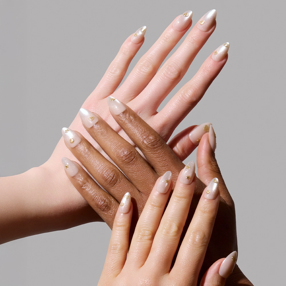 Four hands of varying skin tones display glamnetic’s Sanded Press-On Nail Set, featuring a light nude shade with gold accents and subtle designs. A plain gray background highlights the elegant, beach-inspired manicure. - Product image