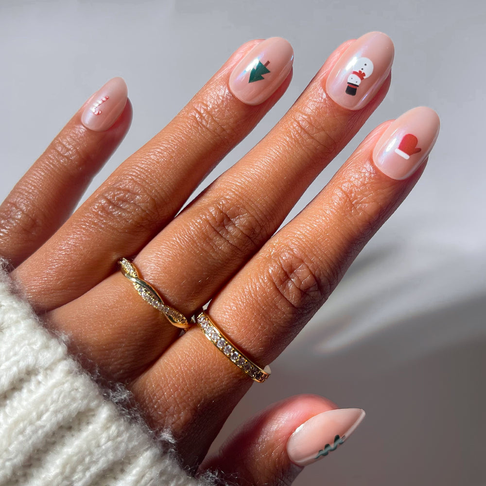 Glamnetic Santa’s Babies oval-shaped press-on nails with a sheer nude base and holiday-themed decals of a candy cane, Christmas tree, snowman, mitten, and garland.