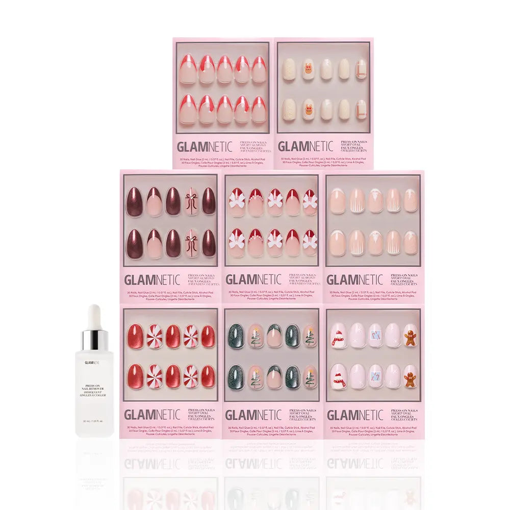 Nine boxes of glamnetic Santa's Workshop festive press-on nails are stacked in a pyramid, featuring red, pink, and holiday designs. A white bottle of cuticle oil with a dropper sits in the bottom left corner. - Product image