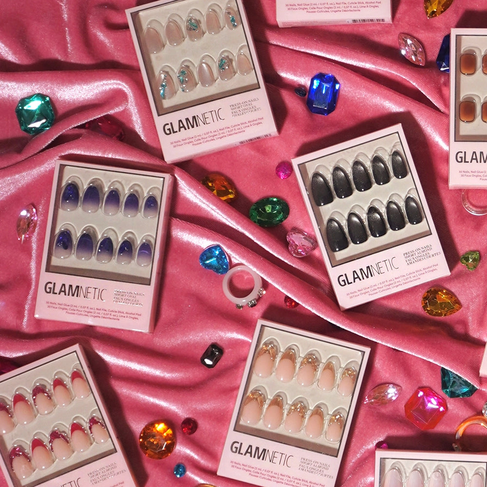 Boxes of glamnetic press-on nails, featuring the Sapphire and other short oval styles in various colors, are displayed on pink satin fabric and accented with vibrant, gem-inspired decorations. - Product image