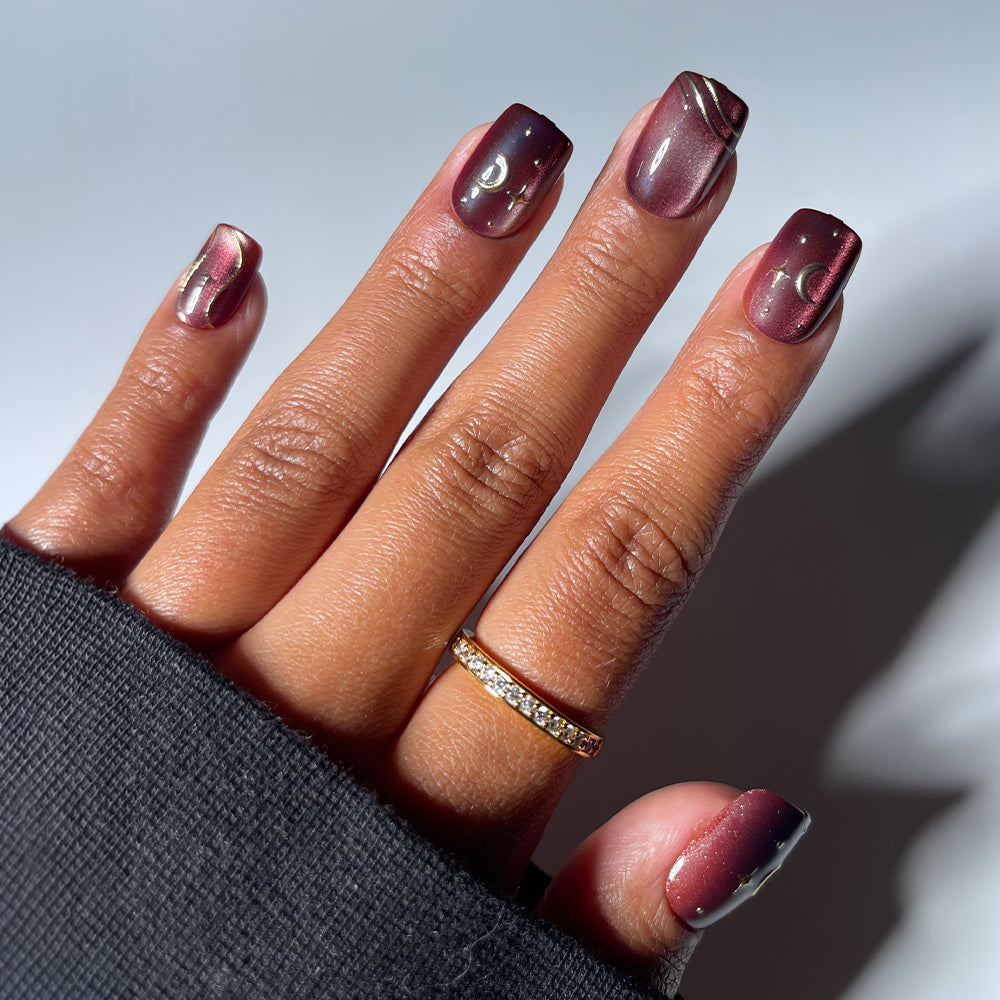 A close-up shot of a hand against a white background, wearing a black long-sleeved garment and featuring glamnetic’s Scarlet Spell press-on nails. The short squoval nails showcase a deep, metallic burgundy cat-eye finish adorned with gold moon and star decals. Easily achieve this striking style with Scarlet Spell for effortless glamour. - Product image