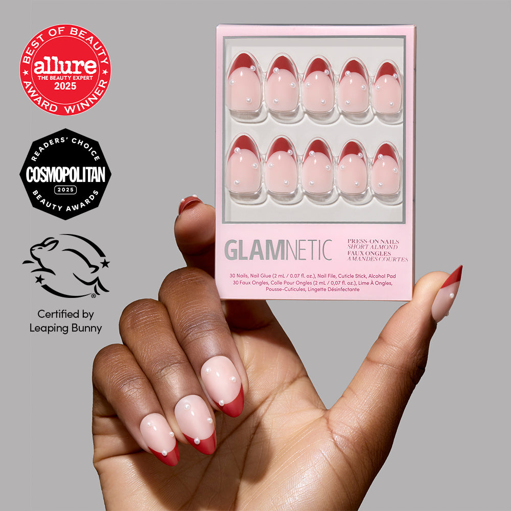 A hand holds a box of Glamnetic Scarlet Pearl press-on nails, which are short, almond-shaped, and feature red French tips with a small white pearl accent on each nail. - Product image