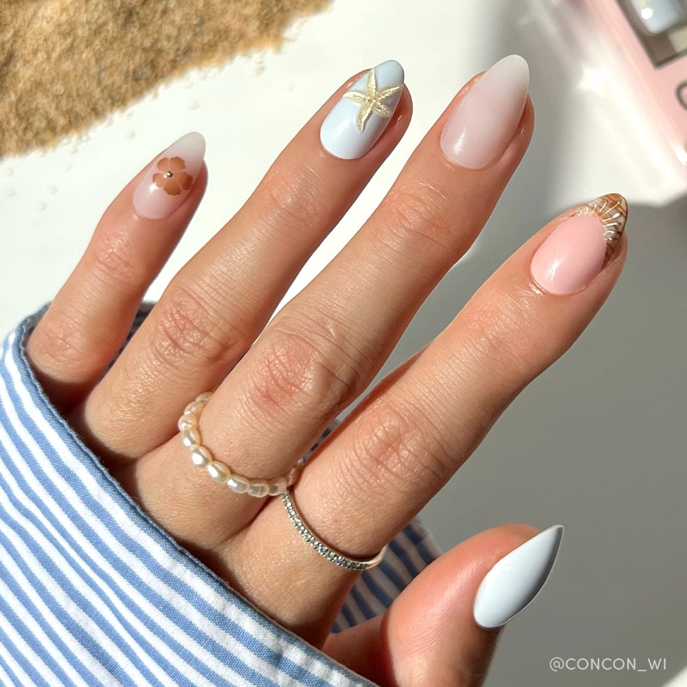 A hand with almond-shaped nails wears glamnetic Sea Star pastel blue and pink nail art with gold seashell, starfish, and 3D shell accents. The person sports pearl and silver rings plus a blue-striped shirt. - Product image