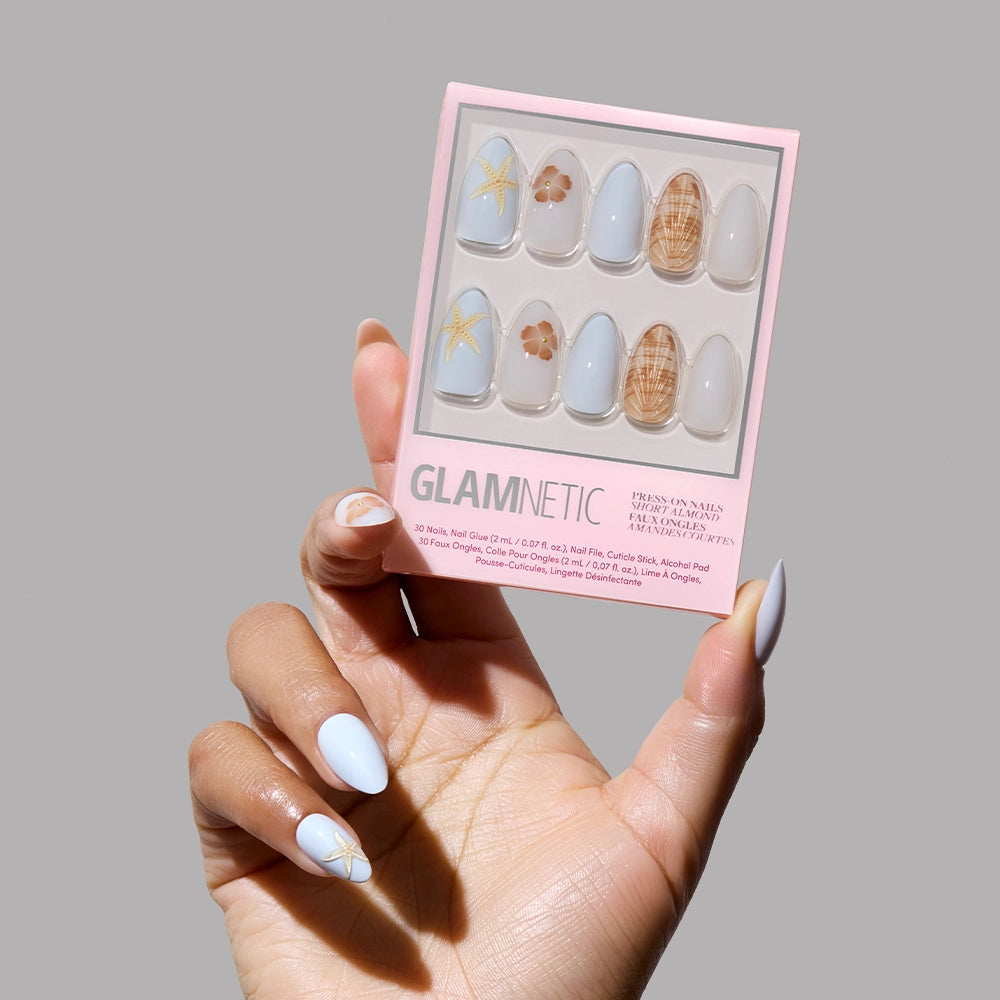 A hand with pastel blue and gold star nail art holds a box of glamnetic Sea Star Press-On Nails, showcasing blue, beige, floral motifs, grid patterns, and 3D shell accents on a plain gray background. - Product image