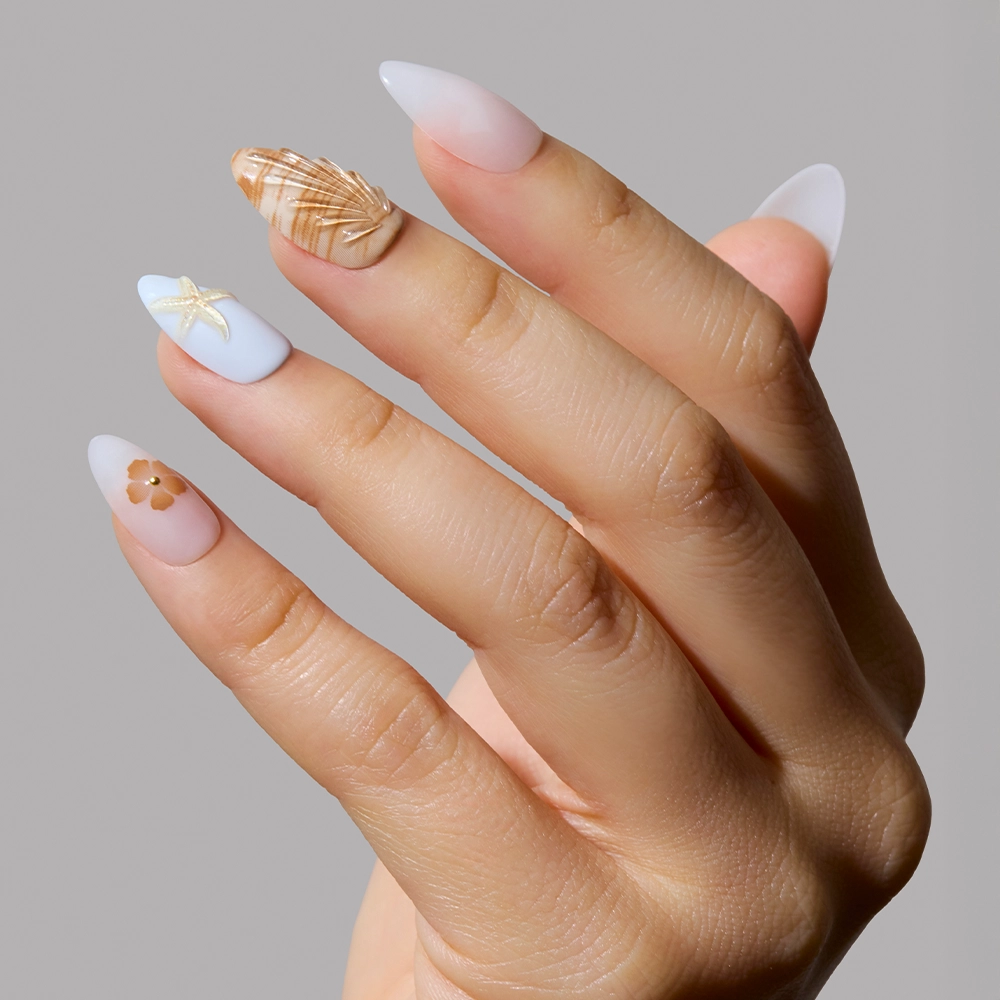 A hand with long, almond-shaped nails shows off the glamnetic Sea Star Press-On Nail Set: soft pastels with 3D shell art on the middle finger, a gold starfish on the index, and floral accents on a neutral base. - Product image