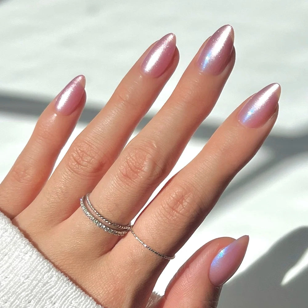 A hand with long, almond-shaped nails painted in glamnetic’s Seaspell—a light pink shade with a shimmery, iridescent sheen and a subtle velvet finish. Thin silver rings adorn the fingers, and a white sleeve is visible.