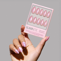 A hand with iridescent pink short almond nails holds a glamnetic Seaspell Press-On Nail Set in a pearly pink, shimmering velvet finish against a neutral gray background.