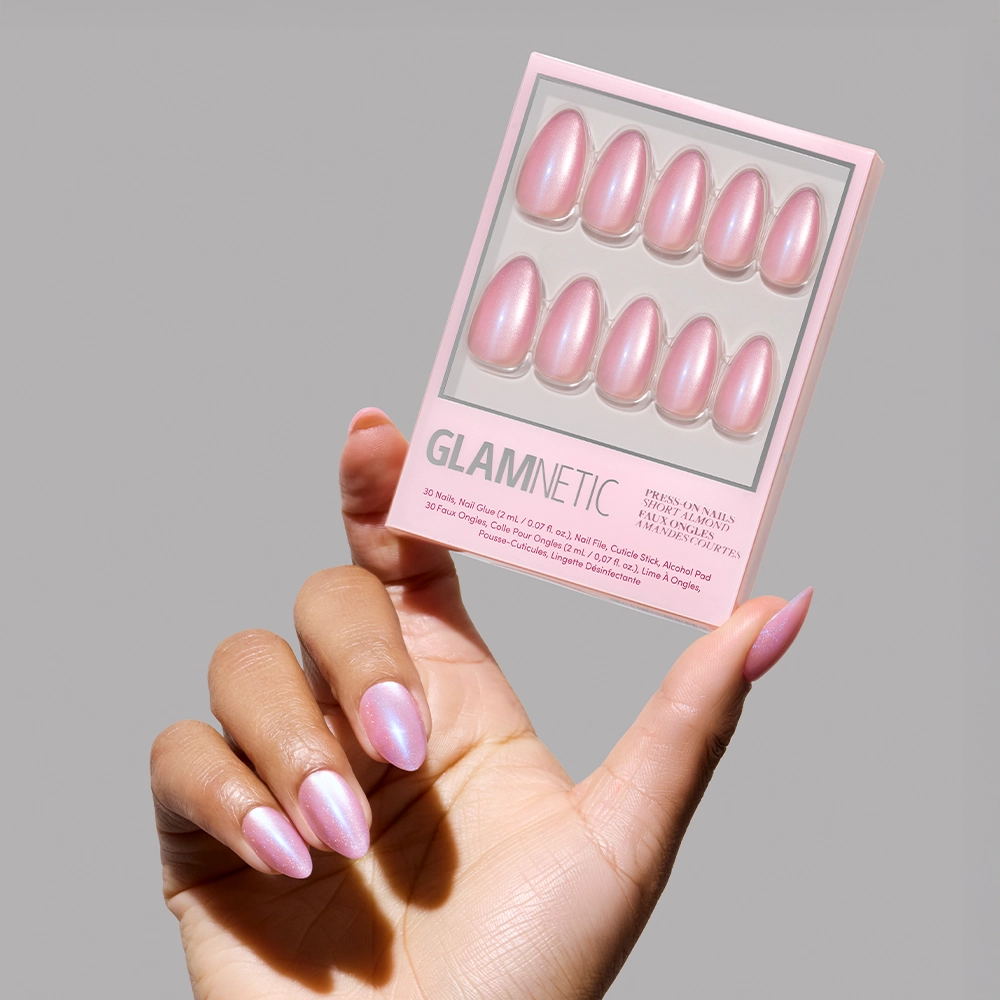 A hand with iridescent pink short almond nails holds a glamnetic Seaspell Press-On Nail Set in a pearly pink, shimmering velvet finish against a neutral gray background. - Product image