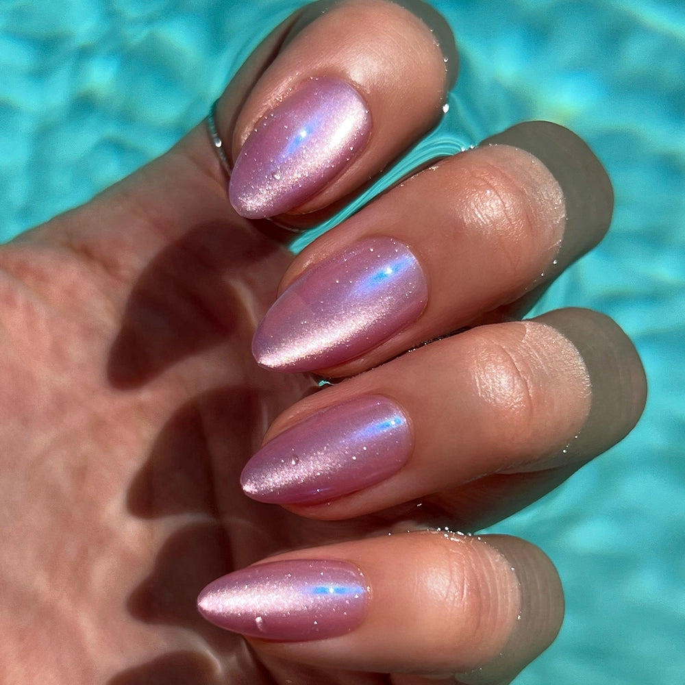 A hand with almond-shaped nails in an iridescent pink velvet finish showcases the glamnetic Seaspell Press-On Nail Set by a clear blue pool, evoking a bright, sunlit outdoor vibe. - Product image