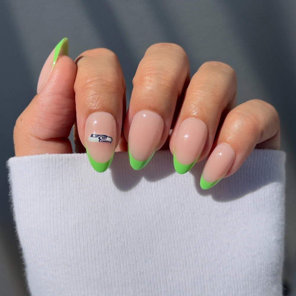 A hand wears the glamnetic Seattle Seahawks Press-On Nail Set, featuring nude almond nails with bright green French tips and a Seahawks logo accent. A white sleeve partially covers the hand—perfect for fans.