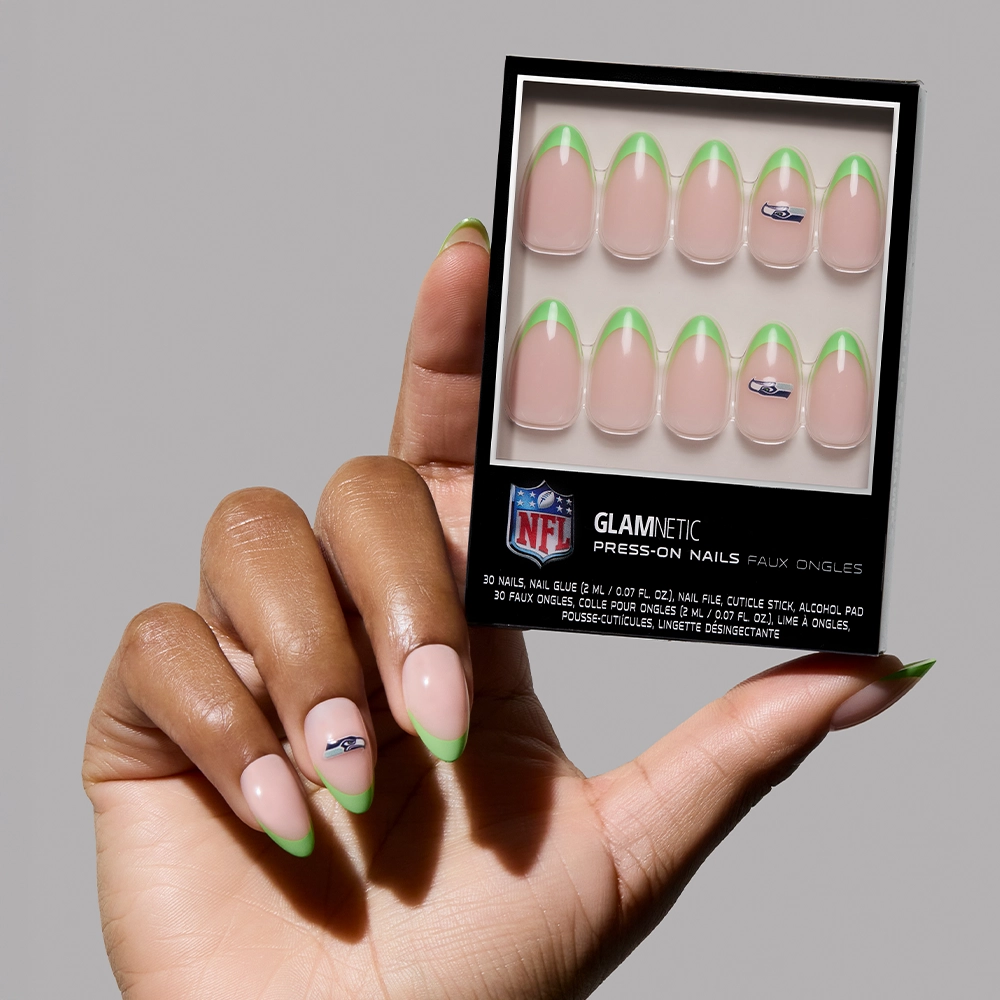 A hand with pale pink nails and green tips holds the Seattle Seahawks Press-On Nail Set by glamnetic, featuring almond-shaped manis and select nails adorned with a blue and silver NFL logo.