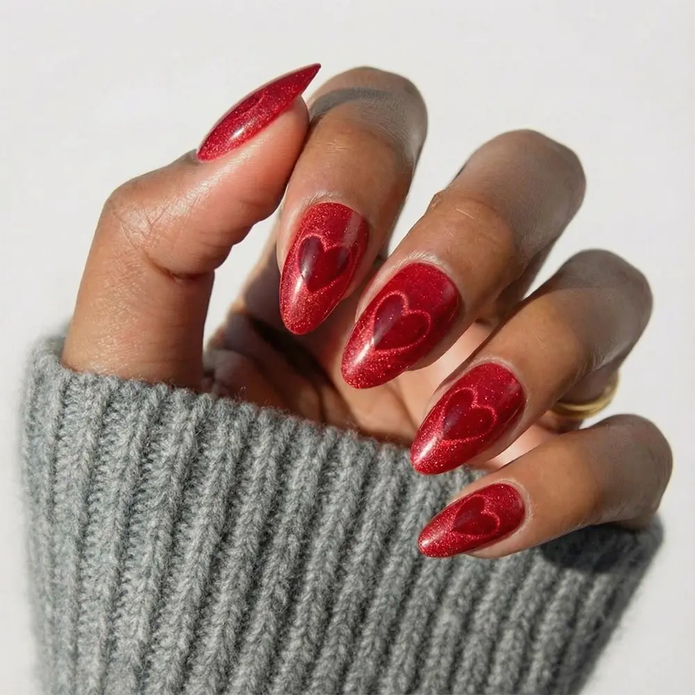 A hand with long, almond-shaped Sent with Love nails by glamnetic, featuring subtle heart designs and a red velvet finish, is shown against a light background. The person wears a gray ribbed sweater sleeve.