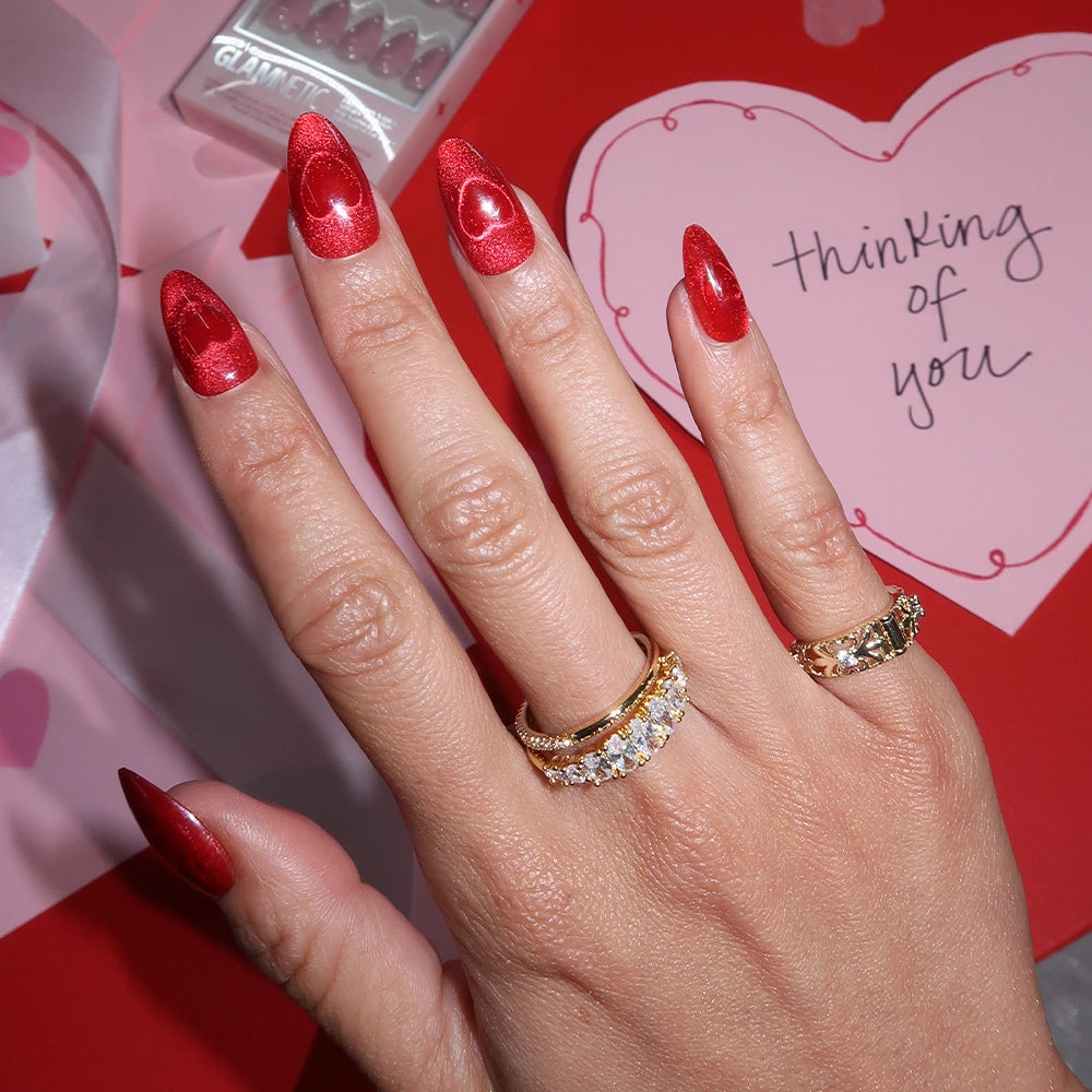 A hand with Sent with Love red velvet heart-patterned nails by glamnetic wears two gold rings with clear stones. In the background is a pink heart card that says thinking of you, on a red surface with ribbon and a Clinique makeup box. - Product image