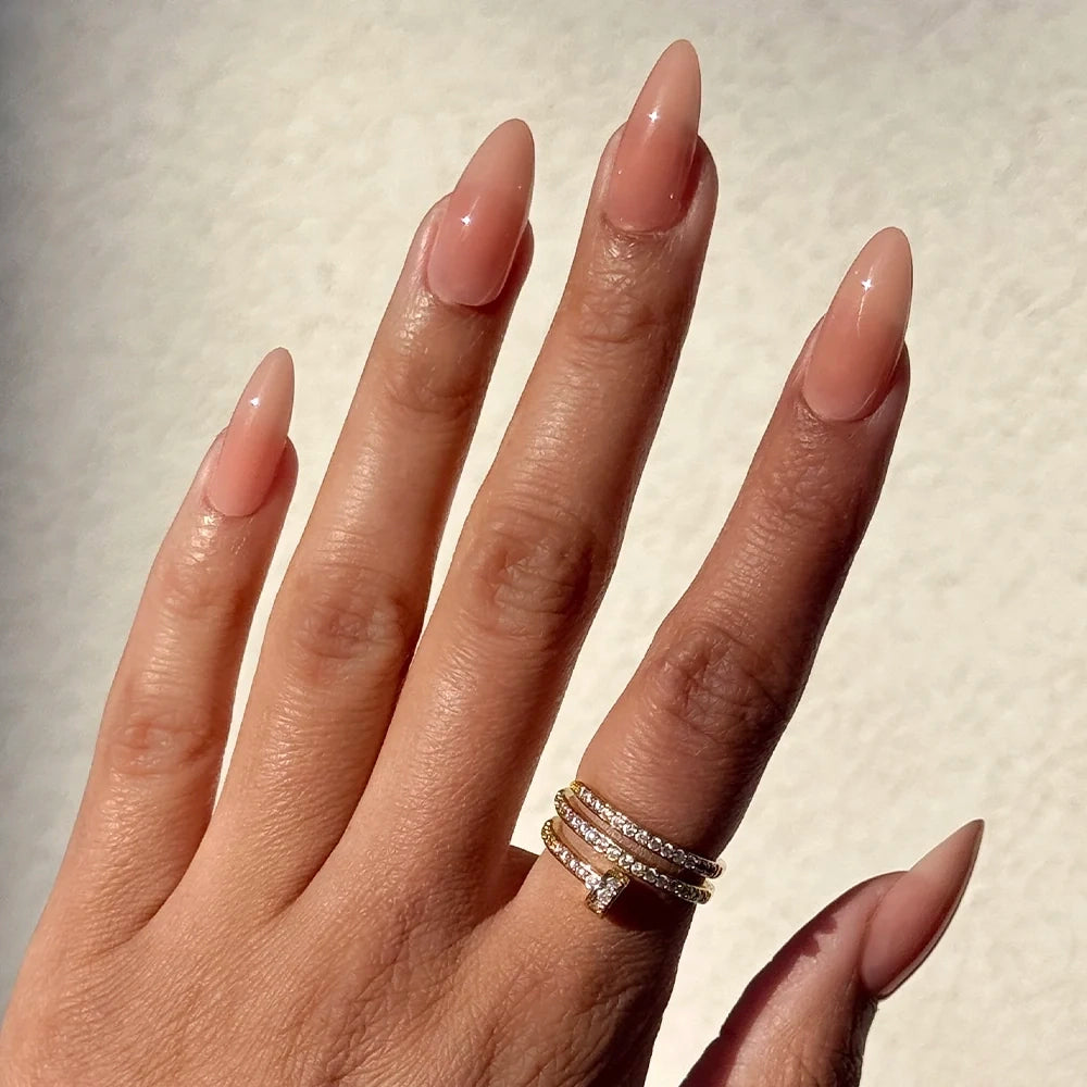 A hand with long, almond-shaped nails in the glossy “Sheer Peach” shade by glamnetic is displayed against a pale background. The ring finger is adorned with three stacked gold rings set with small diamonds. These cruelty-free glamnetic nails elevate the elegant look while maintaining an ethical touch. - Product image
