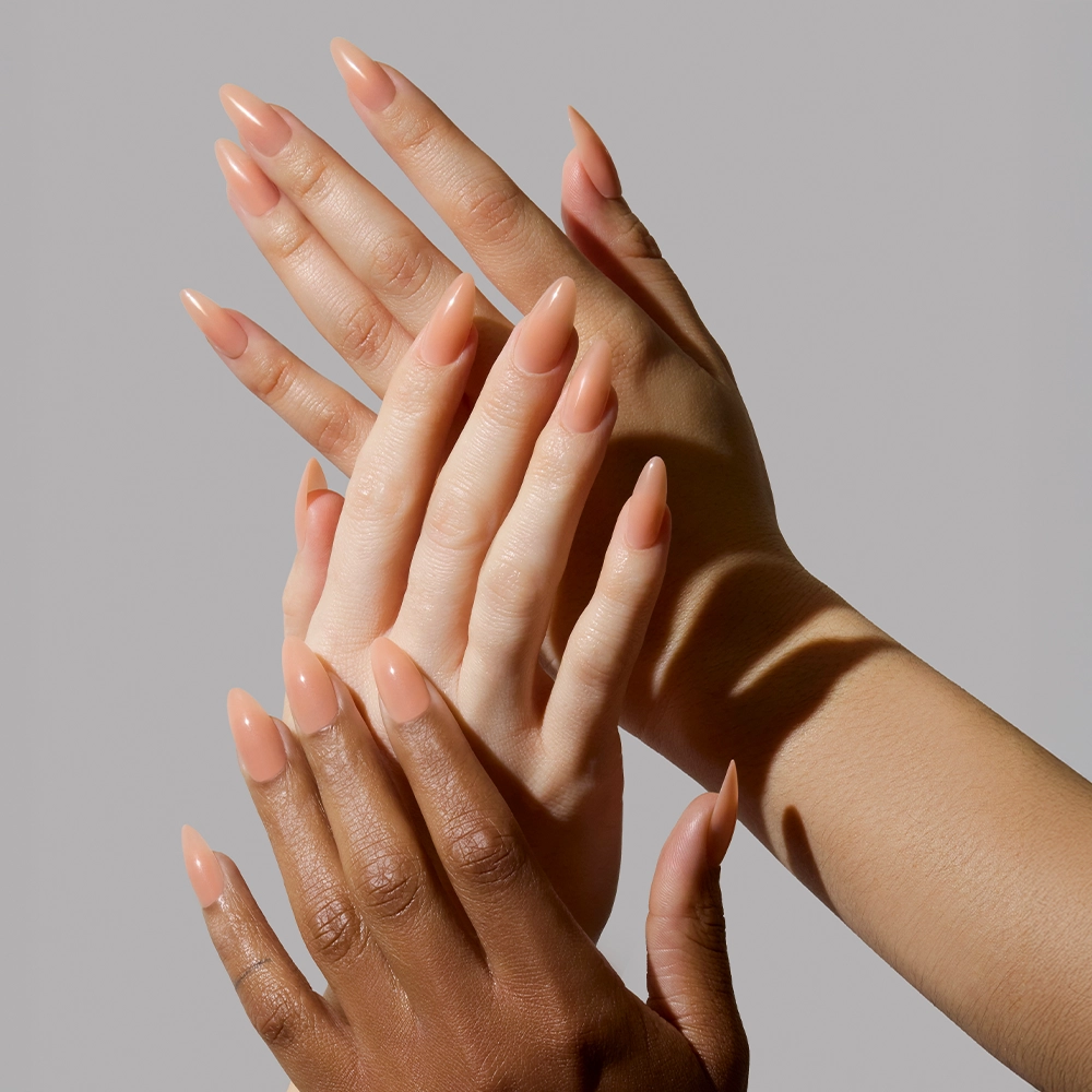 Three hands with different skin tones overlap, showcasing long, almond-shaped nails painted in glamnetic’s Sheer Peach vegan & cruelty-free polish—a semi-transparent peachy nude jelly—against a light grey background. - Product image