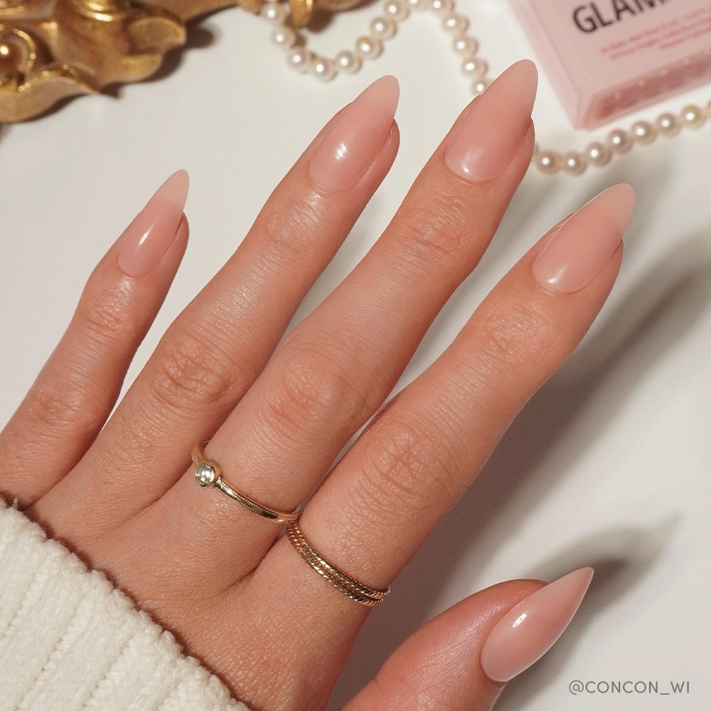 A hand with long, almond-shaped nails in glamnetic’s Sheer Pink shade—a soft-glam staple—wears two gold rings and rests on a white surface beside a gold decor piece, pearls, and a pink box. - Product image