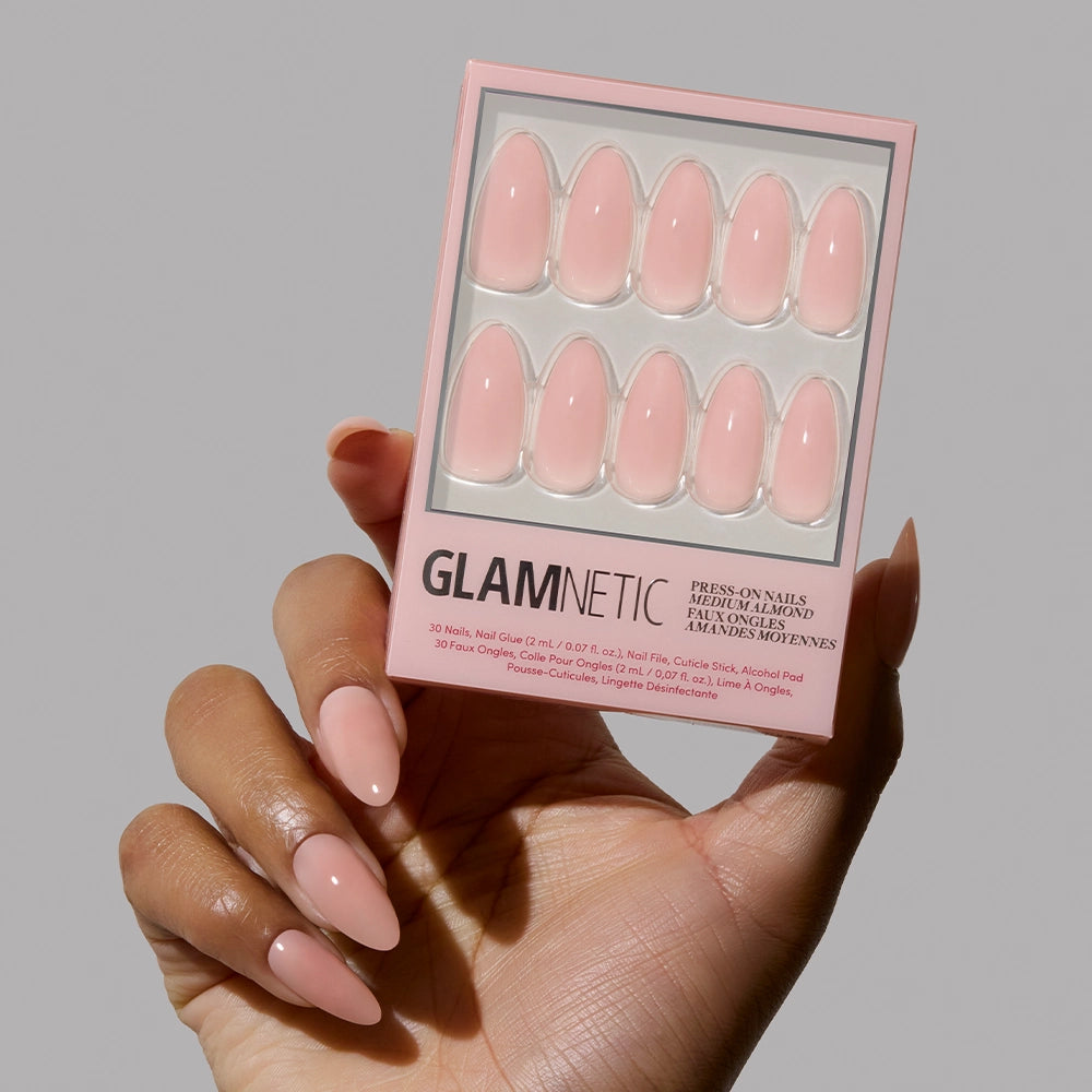 A hand holds a box of glamnetic Sheer Pink press-on nails, while the other shows several applied in the same shade. The vegan, cruelty-free set includes ten glossy, almond-shaped nails on neutral packaging. - Product image