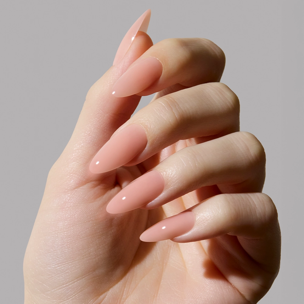 A hand with long, almond-shaped nails painted in glamnetics Sheer Pink polish—a glossy nude pink shade perfect for a clean-girl look—displayed against a plain, light gray background. - Product image