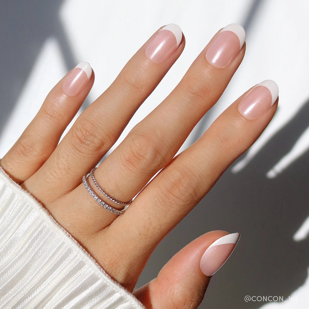 A hand flaunts the glamnetic She’s a Pearl Press-On Nails—a French tip manicure with white tips and a subtle diagonal pinky accent—paired with stacked silver rings and a white ribbed sleeve for an effortlessly chic look. - Product image