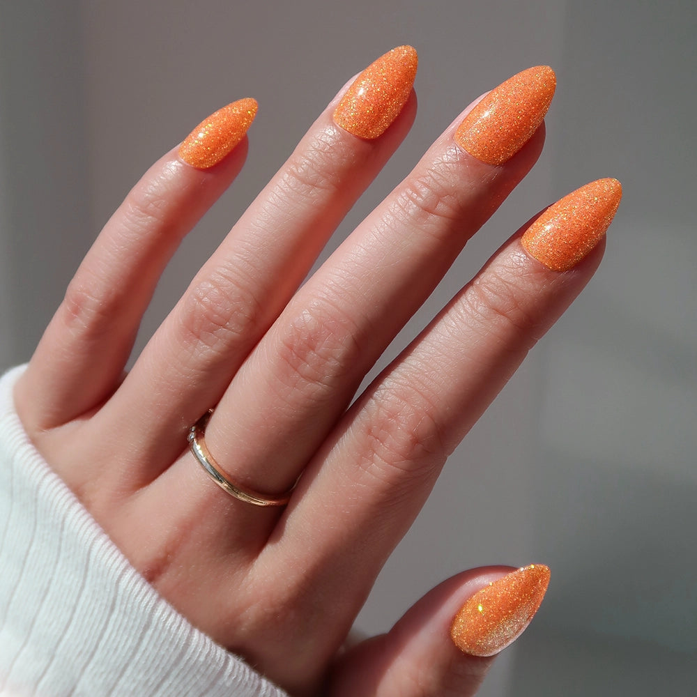 A hand with long, almond-shaped orange nails painted with sparkly polish shows off the glamorous "Showgirl" nail set by glamnetic. The person is wearing a gold ring and a white long-sleeve top. - Product image