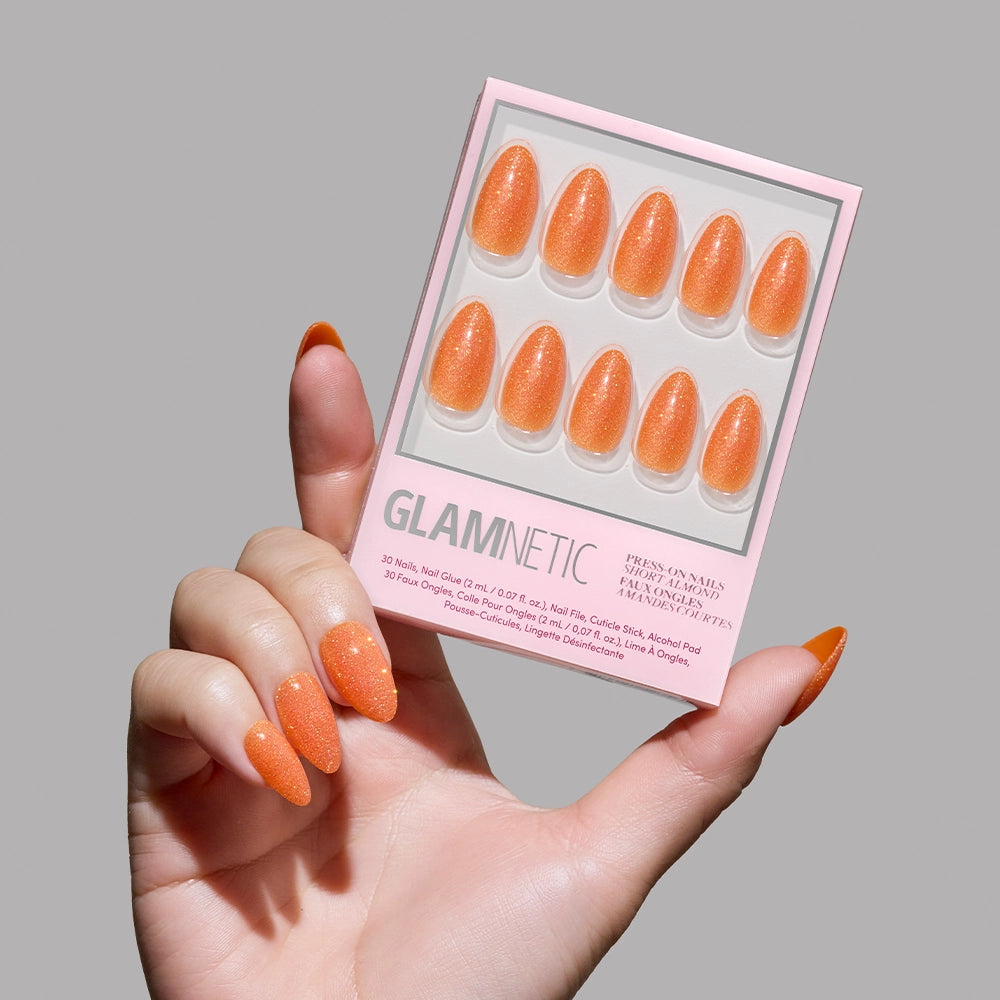 A hand with long, almond orange nails holds a glamnetic Showgirl glitter press-on nail set box against a light gray background, showing ten sparkling orange nails and product details. - Product image