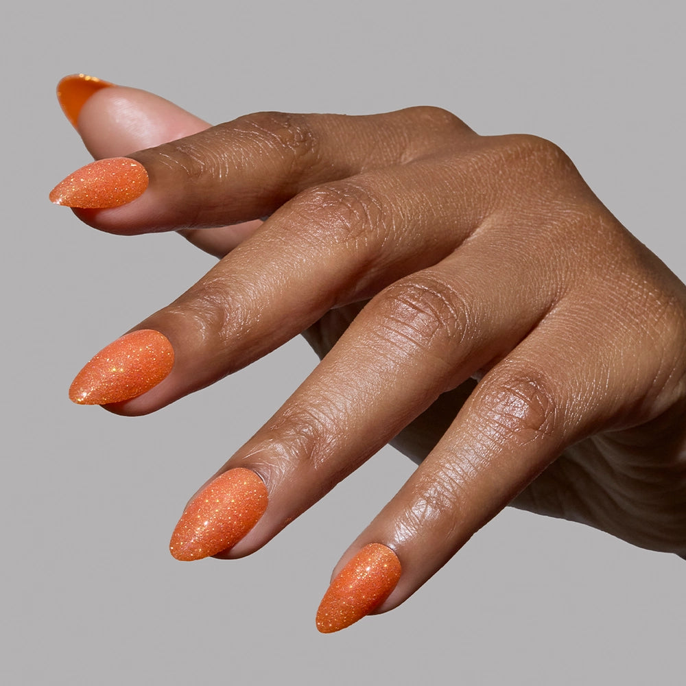 A hand with medium brown skin displays long, almond-shaped orange nails coated in sparkly polish, resembling glamnetic's Showgirl set, against a plain gray background. - Product image