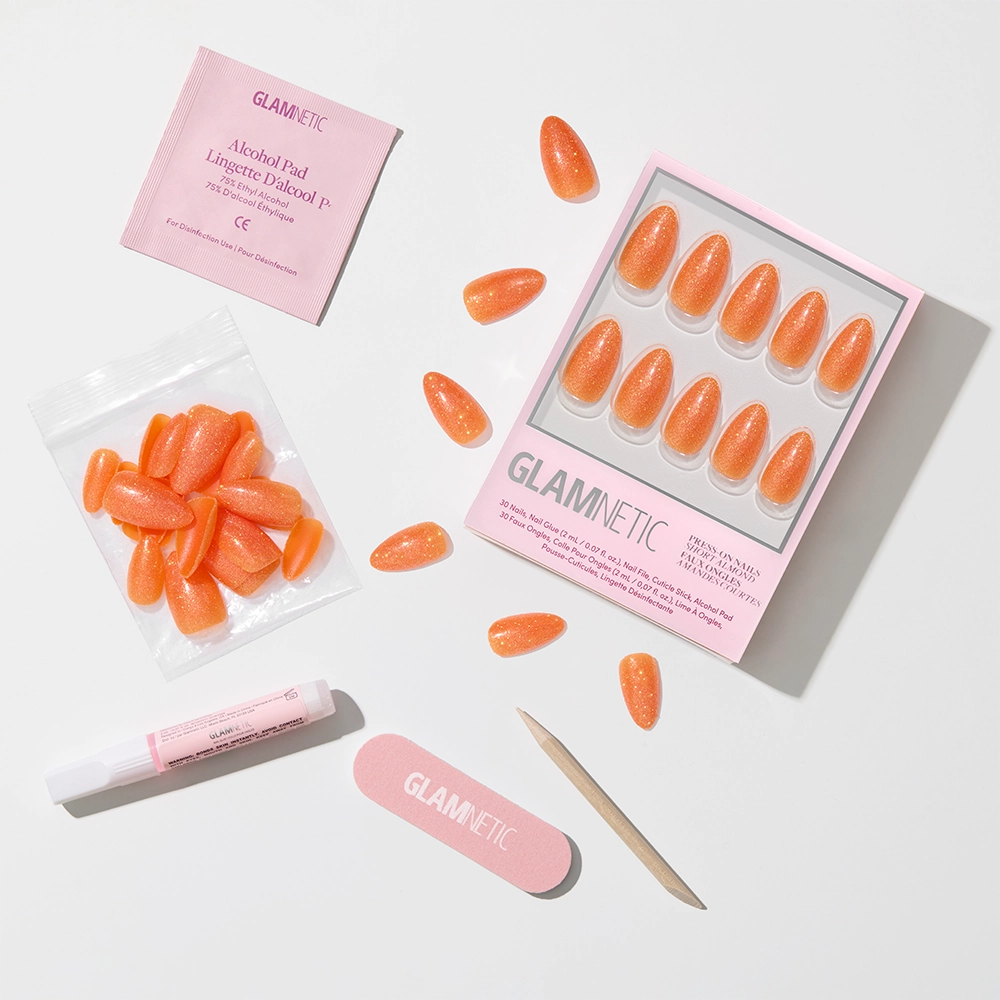 A flat lay displays the Showgirl Nail Set by glamnetic, with almond orange glitter press-on nails, nail file, cuticle stick, glue, alcohol pad, and extra nails in a small bag, all arranged on a white surface. - Product image