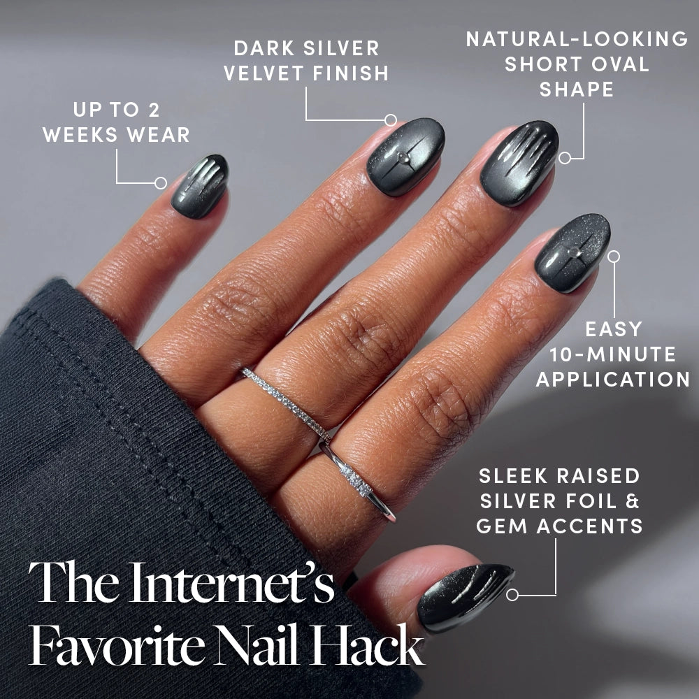A hand with dark silver velvet-finish, short oval jewelry nails featuring silver foil and gem accents. Text overlays highlight features like 2-week wear, 10-minute application, and the title: The Internet’s Favorite Nail Hack. Discover the luxurious look of the Silver Daggers Press-On Nail Set with these trendy velvet press-on nails. - Product image