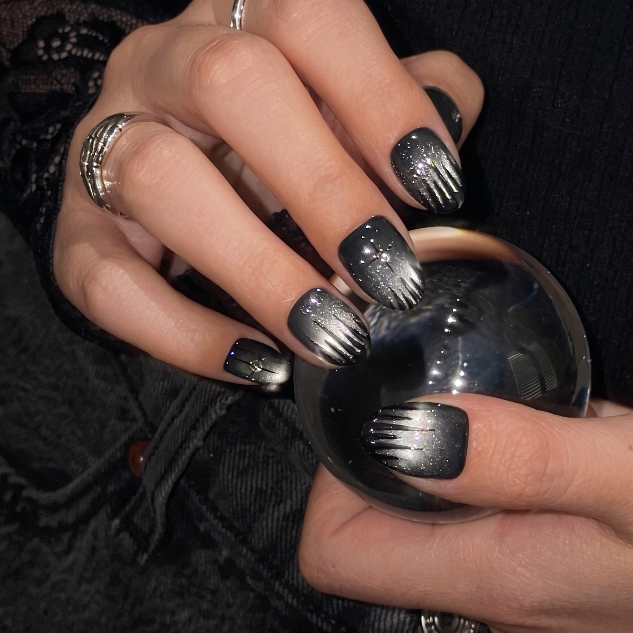 A close-up showcases hands adorned with glamnetic’s Silver Daggers press-on nails—black and silver ombré detailed with glitter and star accents—holding a reflective glass sphere. Silver rings emphasize the statement jewelry nails, complemented by a dark, textured outfit. - Product image