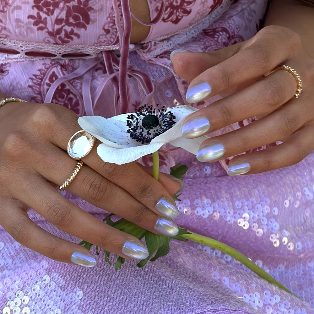 A person with short oval Silver Siren nails by glamnetic, featuring an iridescent blue chrome finish, holds a white flower with a dark center. They wear gold rings and a pale pink, floral sequined dress. - Product image