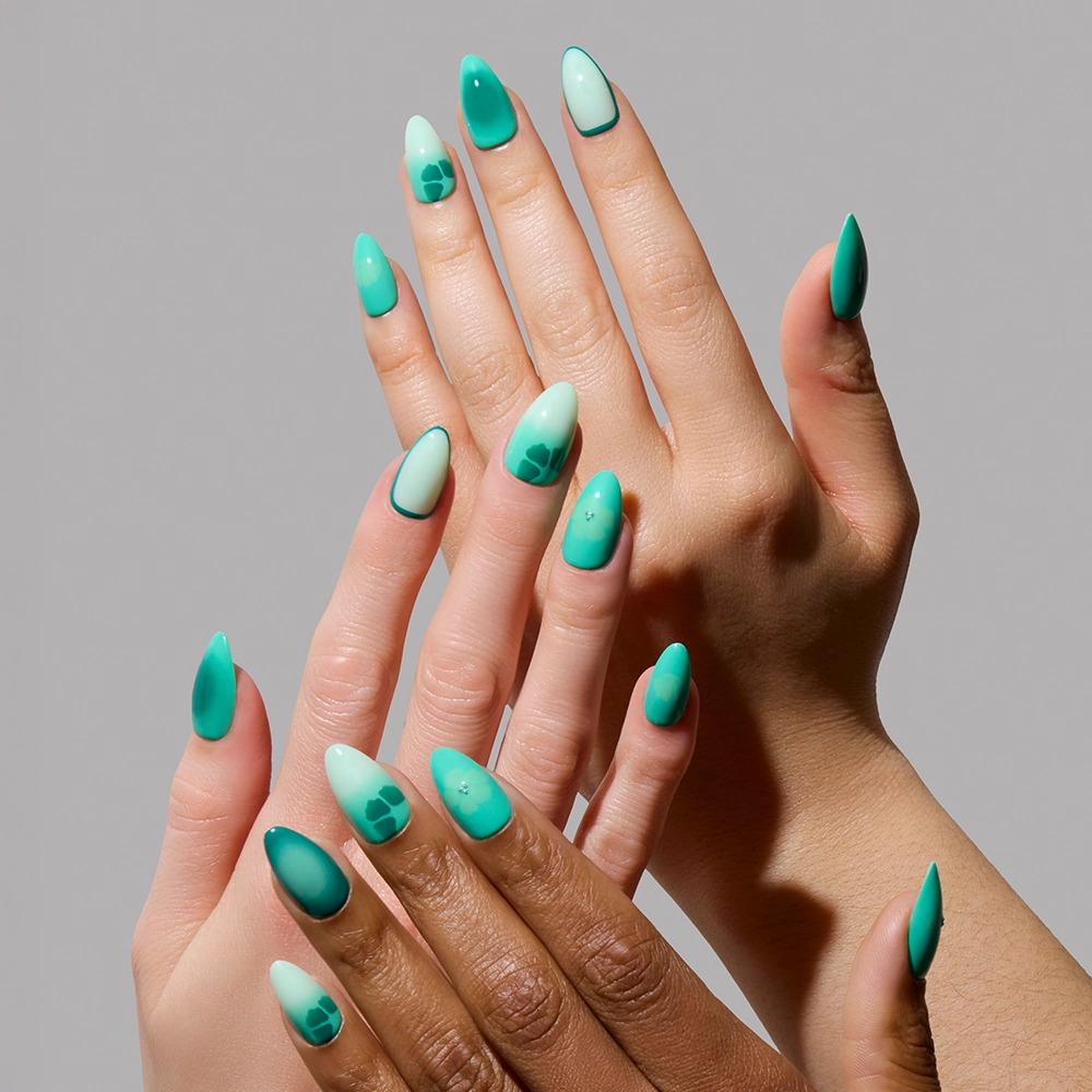 Four hands display glamnetic’s “Siren Green” nails in long, almond shapes, painted teal and mint with geometric patterns and a glossy aquamarine gradient—perfect for mermaid-inspired glam—set against a neutral gray background. - Product image