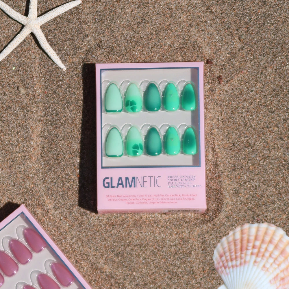 A box of glamnetic Siren Green press-on nails in green rests on a sandy beach with seashells and a starfish, offering mermaid-inspired glam. A pink set peeks from the corner. - Product image