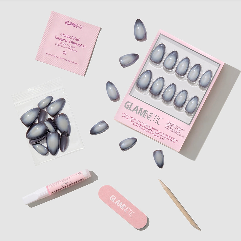 The glamnetic Smoke N’ Mirrors set includes salon-quality gray press-on nails, pink nail glue, a nail file, wooden cuticle stick, alcohol pad, and extra nails—all arranged neatly on a light gray background. - Product image