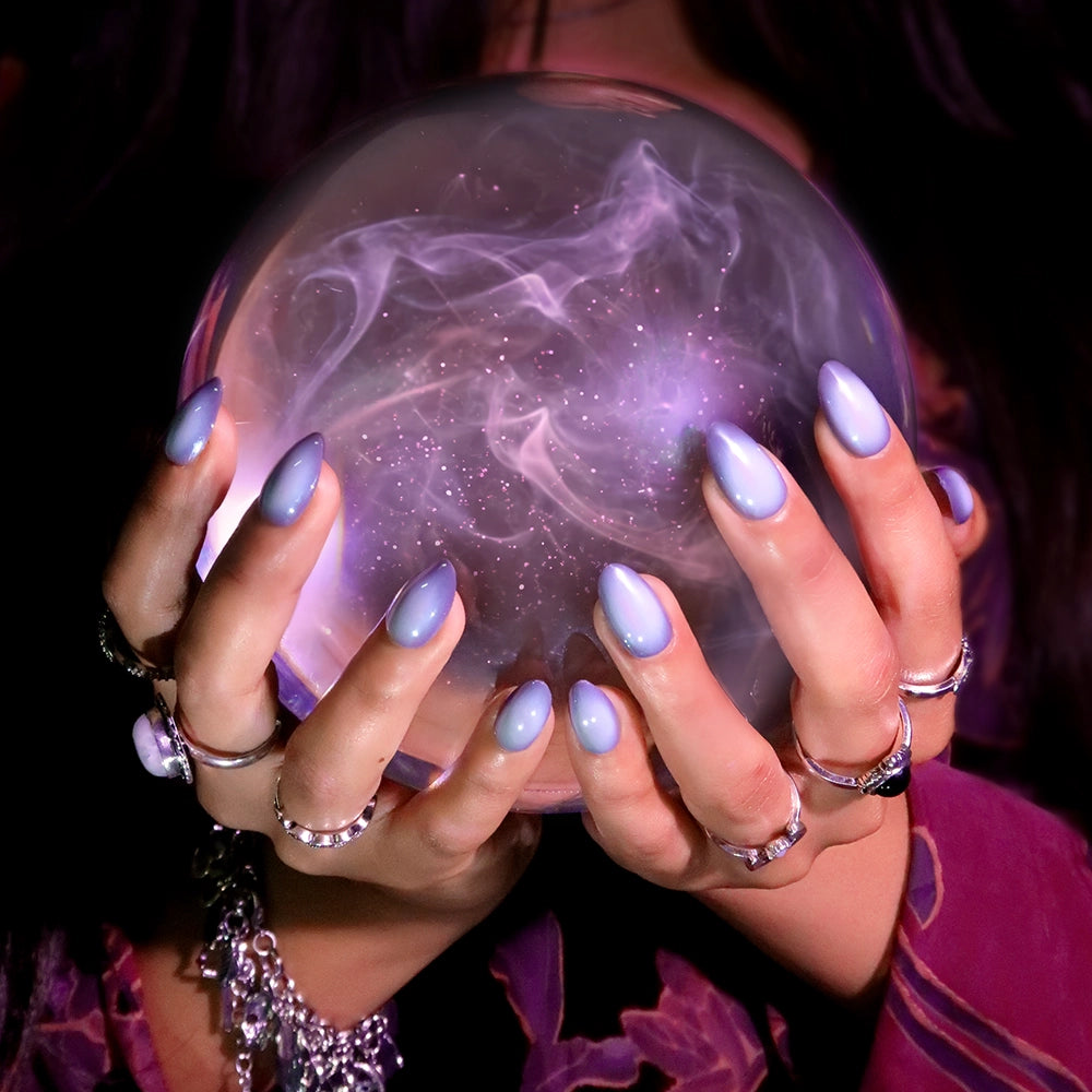 A person with short almond nails painted in the glamnetic Smoke N’ Mirrors lavender hue and adorned with silver rings holds a glowing, smoky crystal ball, evoking themes of fortune telling and mysticism. The salon-quality durability of the Smoke N’ Mirrors press-on nails enhances the scene’s elegance and mystery. - Product image