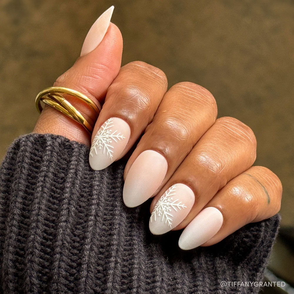 A hand with almond-shaped, matte nude nails from the glamnetic Snowdrift set features raised snowflake accents on two accent nails. The look is styled with a chunky gold ring and a dark ribbed sweater for added chic appeal. - Product image