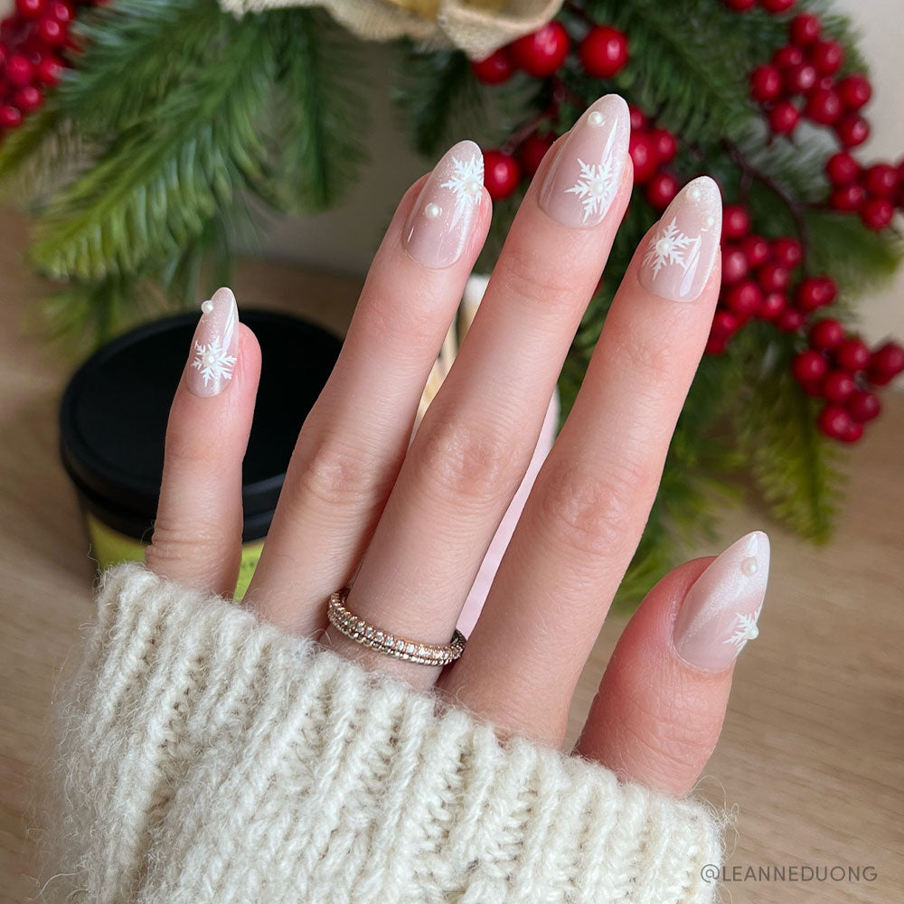 A hand with short almond nails wears glamnetic’s Snowfall, featuring a sheer pink base, snowflake designs, and white dots. The person is in a cream ribbed sweater with a gold ring; festive greenery and red berries appear in the background. - Product image