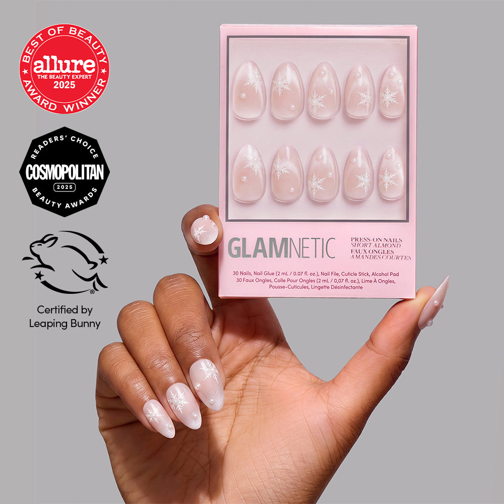 A hand with long, almond-shaped nails featuring white floral designs holds a box of the glamnetic Snowfall press-on nail set. Beauty award logos and a Certified by Leaping Bunny cruelty-free symbol are displayed on the gray background, emphasizing these salon-quality press-on nails with an elegant velvet finish. - Product image
