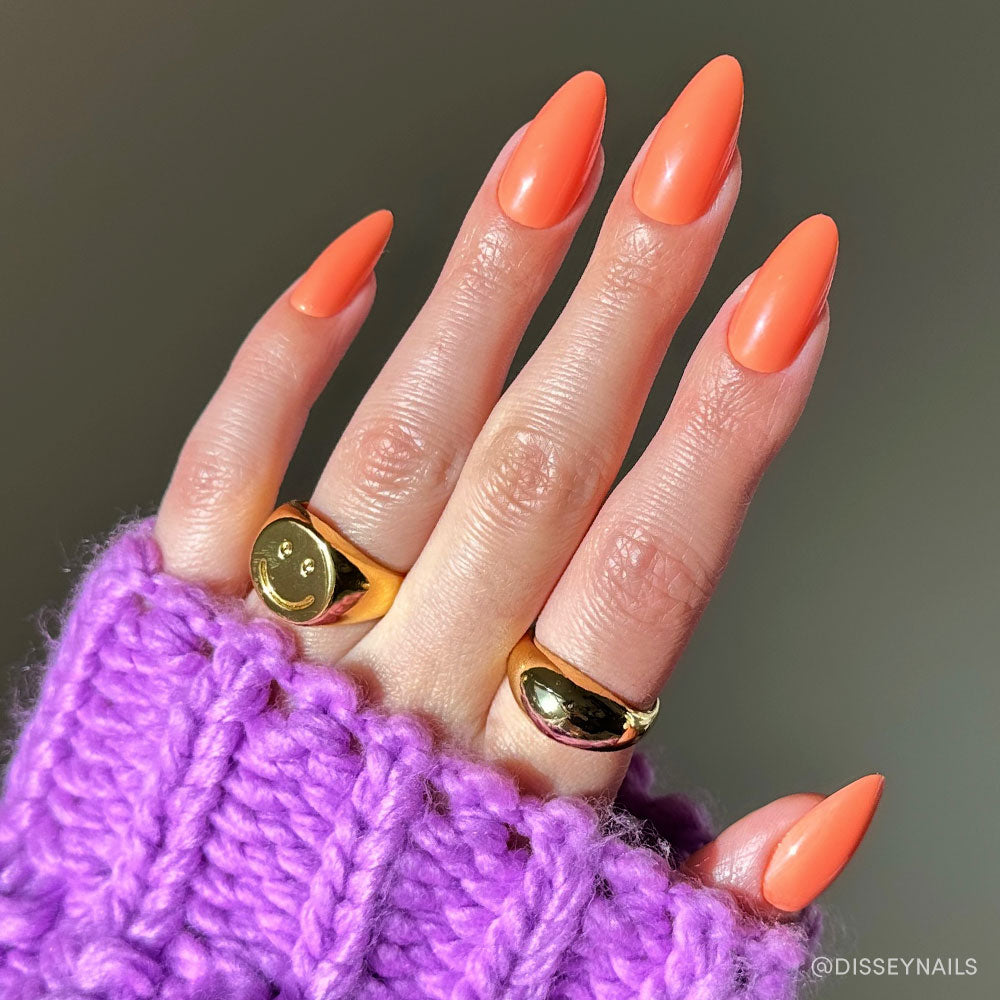 A hand with long, almond-shaped nails painted in Soft Tangerine by glamnetic, adorned with two gold rings—one featuring a smiley face—rests on a chunky, purple knitted sweater. - Product image