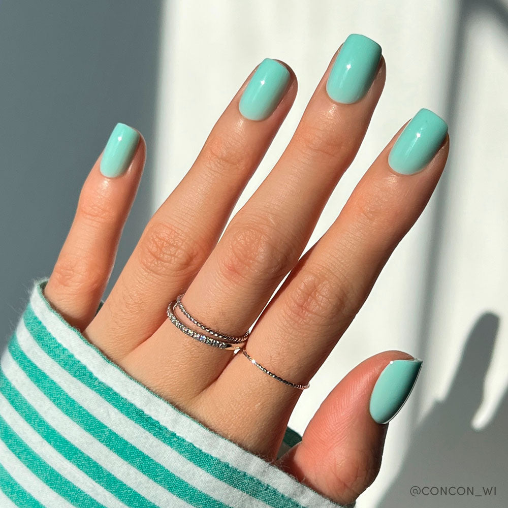 A hand with neatly manicured short squoval nails painted in glamnetics Soft Teal. Thin silver rings adorn the fingers, and a green-and-white striped sleeve partially covers the wrist as bright sunlight casts soft shadows. - Product image