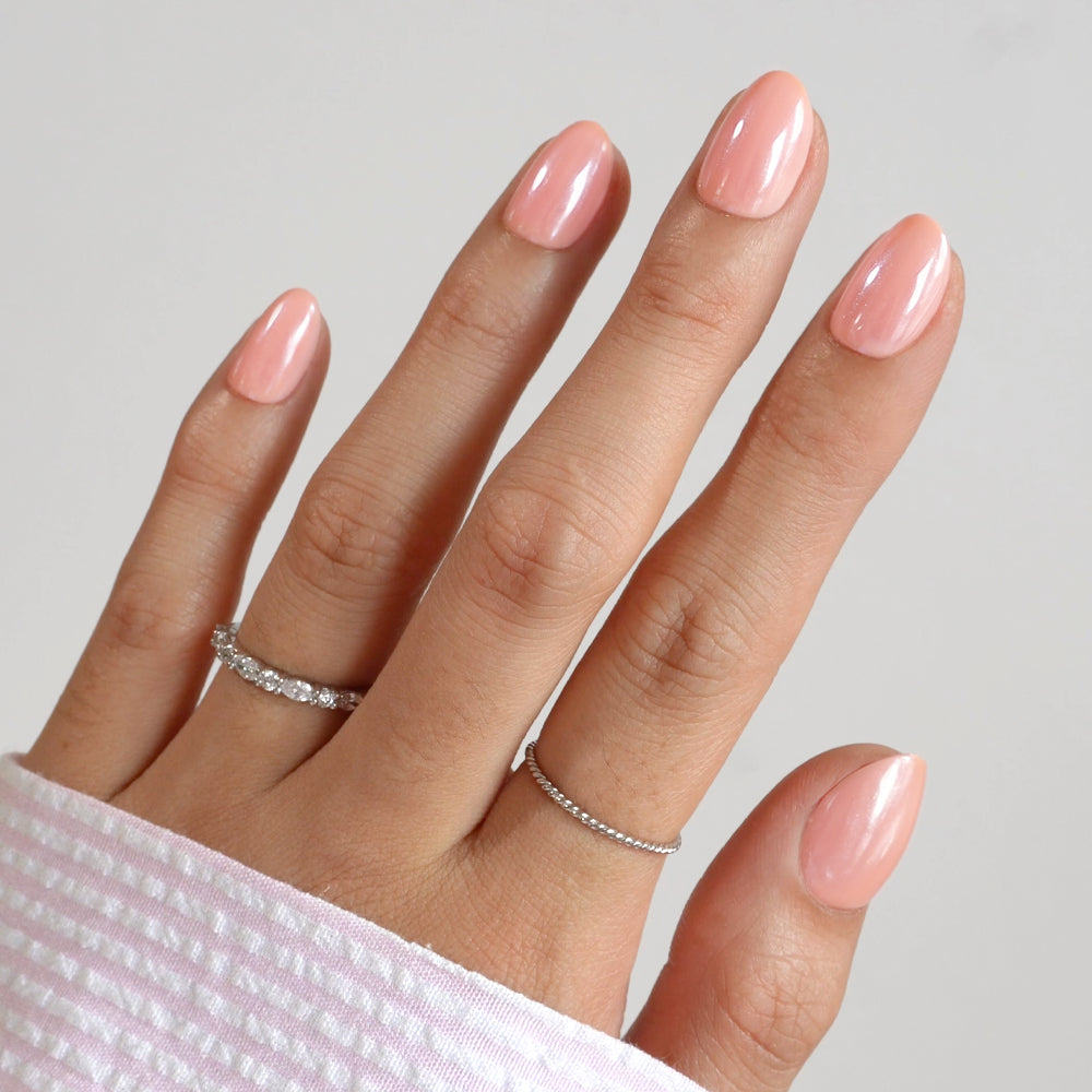 A hand with glossy, light pink "Soft Skin" press-on nails by glamnetic in a short oval shape—wearing two silver rings and partially covered by a pink and white striped sleeve—set against a plain background. - Product image