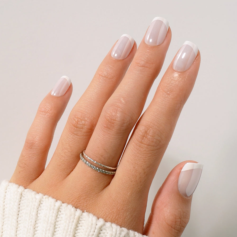 A hand adorned with glamnetic's Soft Tulle French manicure—showcasing classic French tips on short squoval nails—and two thin silver rings, rests on a white knit sweater. - Product image