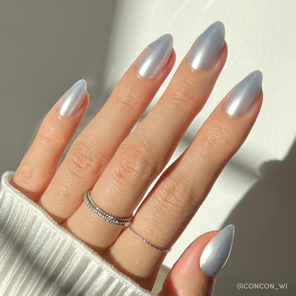 A hand with salon-quality, long almond-shaped nails in glamnetic’s Something Blue shade. The nails are painted in a shiny metallic blue, paired with three delicate rings and a white ribbed sweater sleeve partially covering the wrist. - Product image
