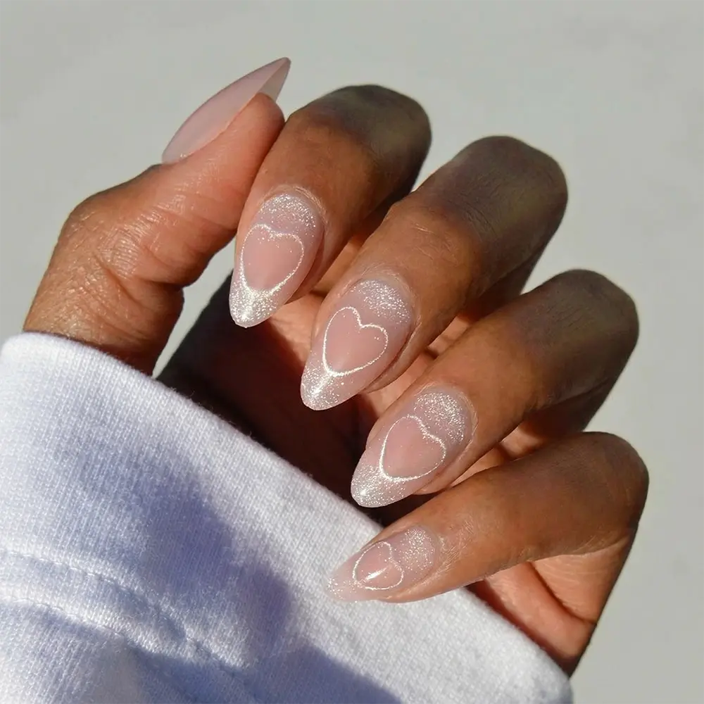 A hand displays glamnetic's "Soulmate Energy" almond-shaped velvet press-on nails in shimmery nude, each adorned with delicate white heart designs. The person is wearing a white long-sleeve shirt.