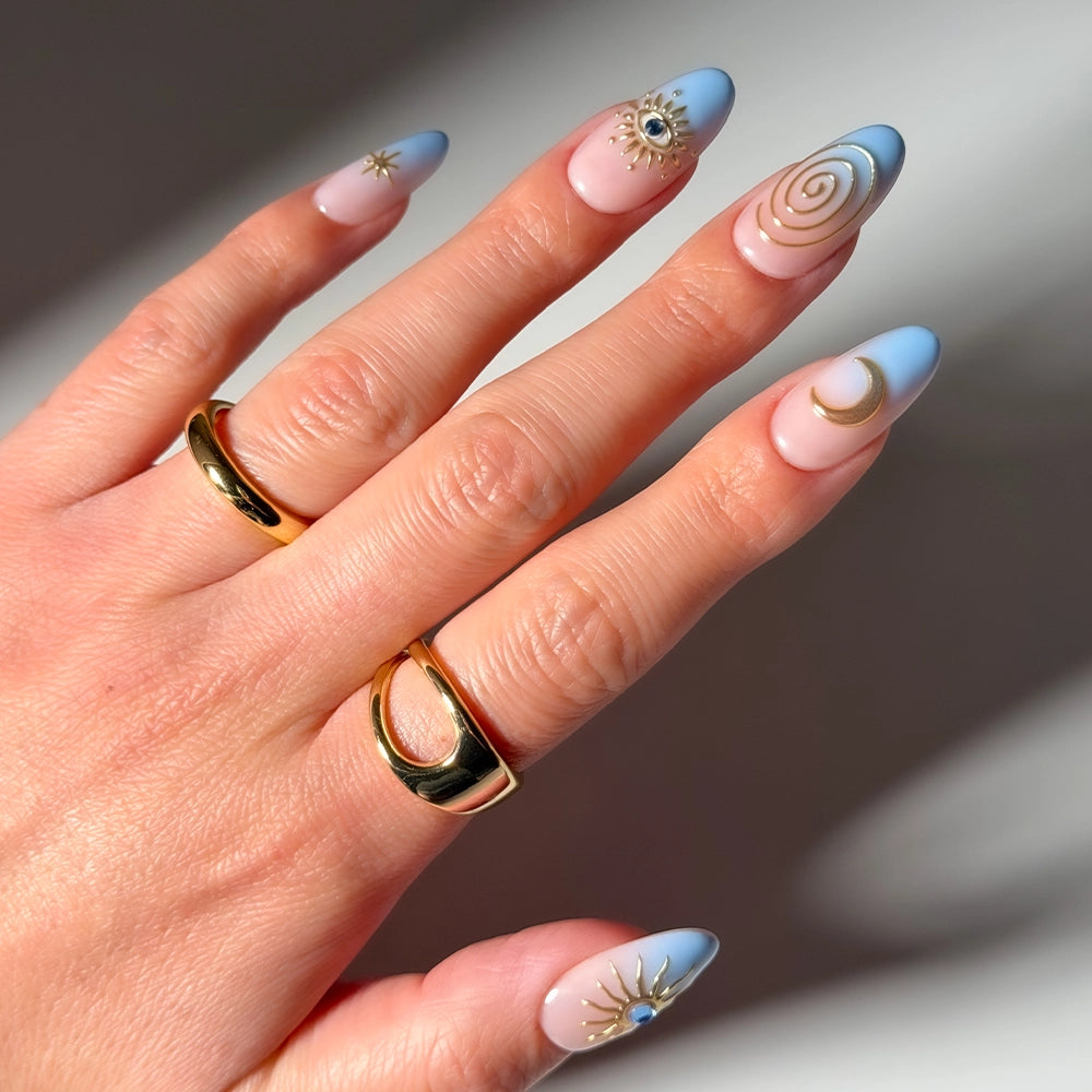 Spirited Sky by glamnetic features blue ombre French tips and nude stiletto nails with gold suns, eyes, spirals, and moons. Two gold rings finish the look in sunlight—perfect press-on nails for a cosmic vibe.