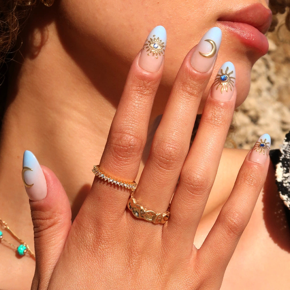 A hand flaunts glamnetic's Spirited Sky nails—long, almond-shaped with blue ombre French tips and gold celestial designs. Gold rings and jewelry adorn the person, whose curly hair and partial face appear in the background. - Product image