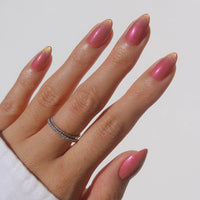 A hand with short almond nails features glamnetic Starbeam, a shimmery pink polish with a gold sheen and velvet finish. Two thin stacked rings decorate the ring finger, paired with a white sleeve for a cruelty-free manicure.