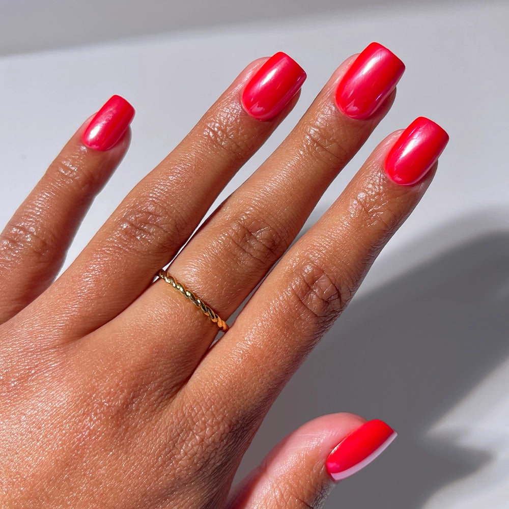 A hand with medium brown skin wears a thin gold twisted ring, showcasing Glamnetic’s Statement Red salon-quality press-on nails in a shiny bright red polish. Nails are neatly manicured against a plain white background.