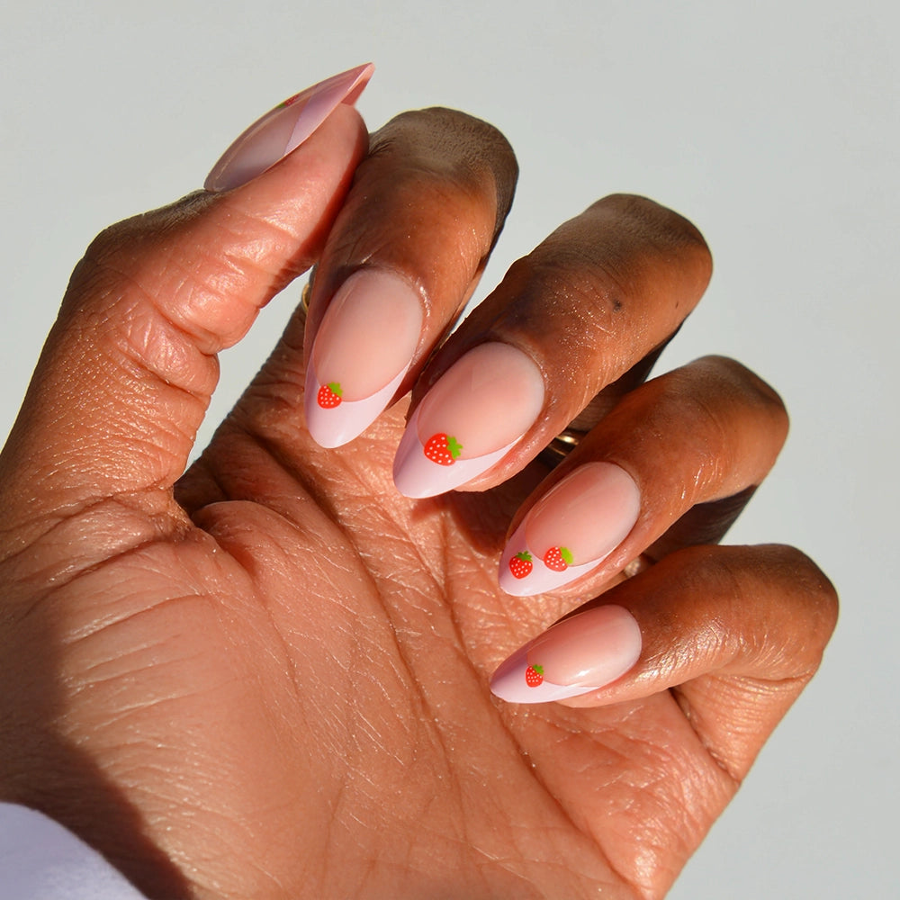 A hand with almond nails painted in a nude pink shade with white tips, each nail adorned near the cuticle with glamnetic's Strawberry Bits cute strawberry nail art.