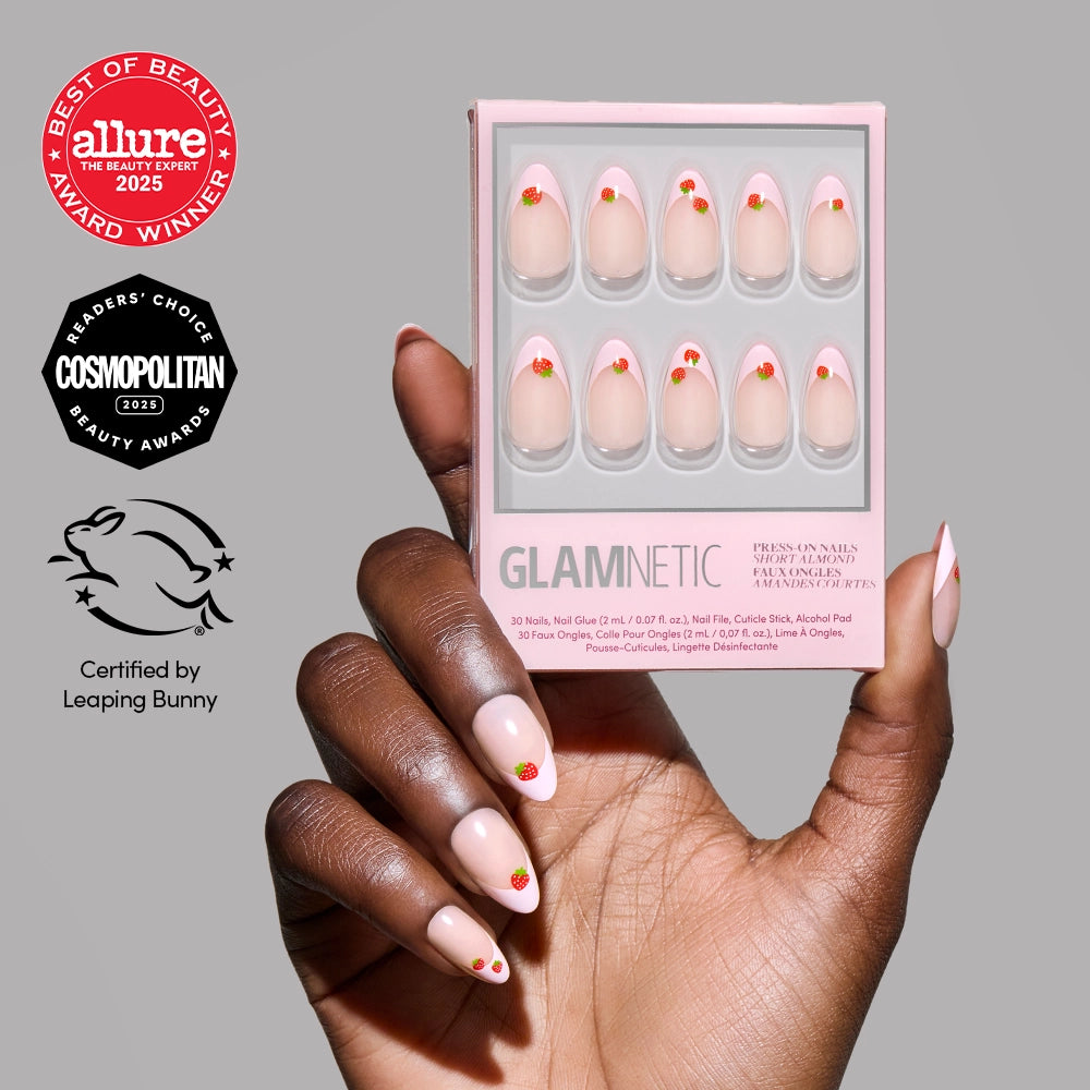 A hand with light pink almond press-on nails decorated with small red hearts and strawberry art holds a box of glamnetic Strawberry Bits nails. Beauty award badges and a cruelty-free logo are shown on the grey background.