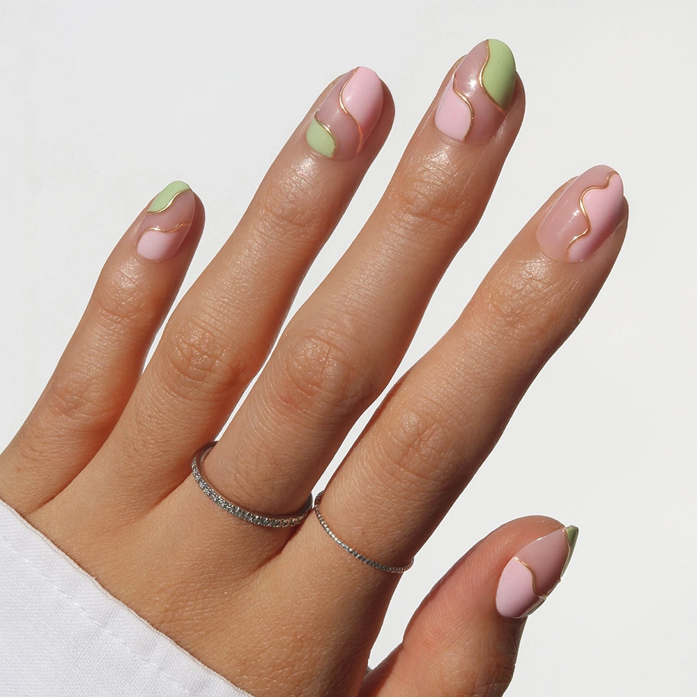 A hand with almond-shaped café-cute nails features glamnetic's Strawberry Matcha press-ons, showcasing pastel pink and green wavy designs separated by thin gold lines, paired with two simple silver rings and a white sleeve.